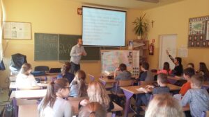 Sala szkolna, uczniowie siedzą w ławkach i aktywnie uczestniczą w zajęciach prowadzonych przez nauczyciela